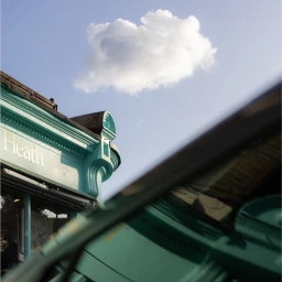 Close-up architectural shot of a teal shop facade and a single white cloud in a blue sky by Nico Froehlich.
