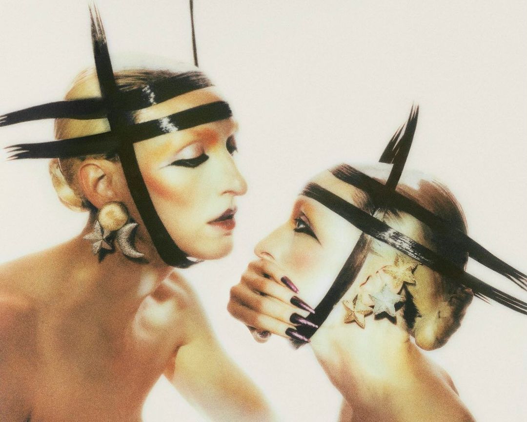 Two models with makeup and avant-garde headwear, posing in a studio. The image showcases unique fashion and artistic makeup.