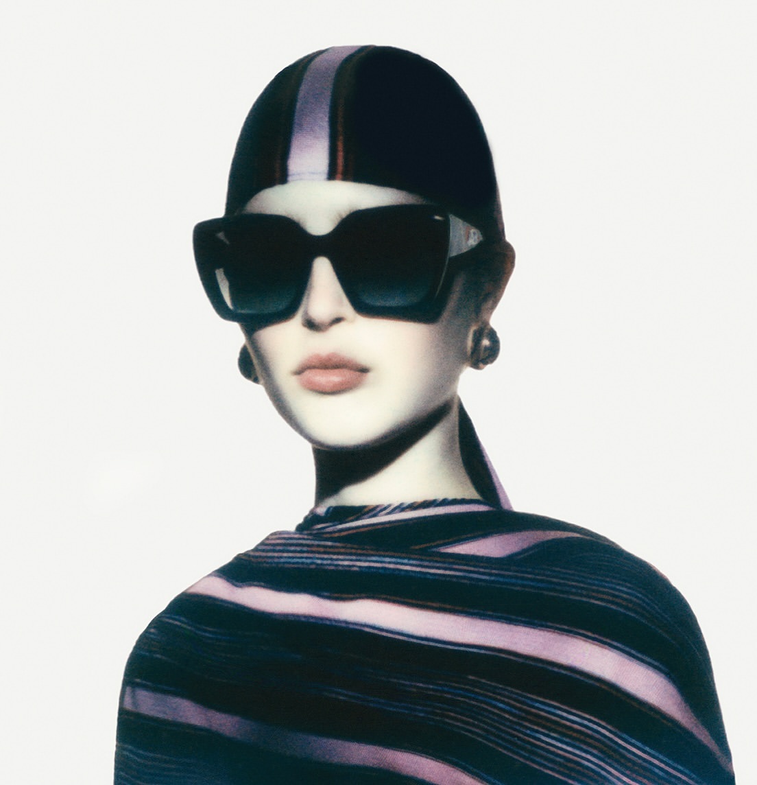 Portrait of an adult female fashion model wearing sunglasses, a headscarf, and striped clothing against a bright white background.