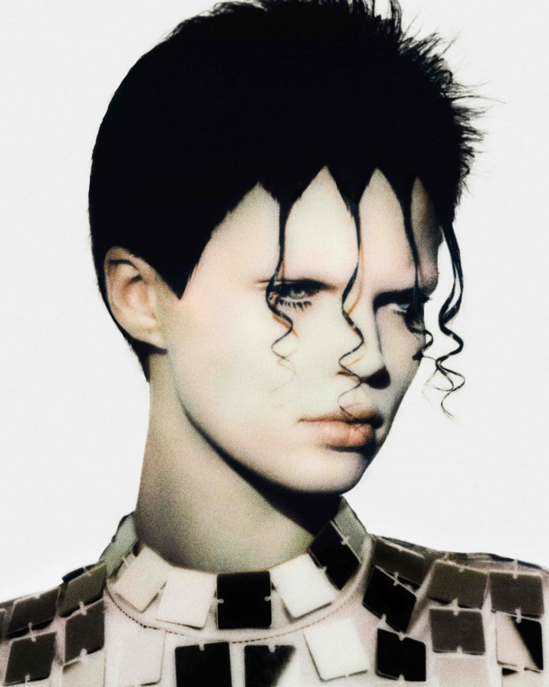 Close-up portrait of a person with stylized black geometric hair and pale skin, wearing a mirrored square-patterned garment in a photo studio.