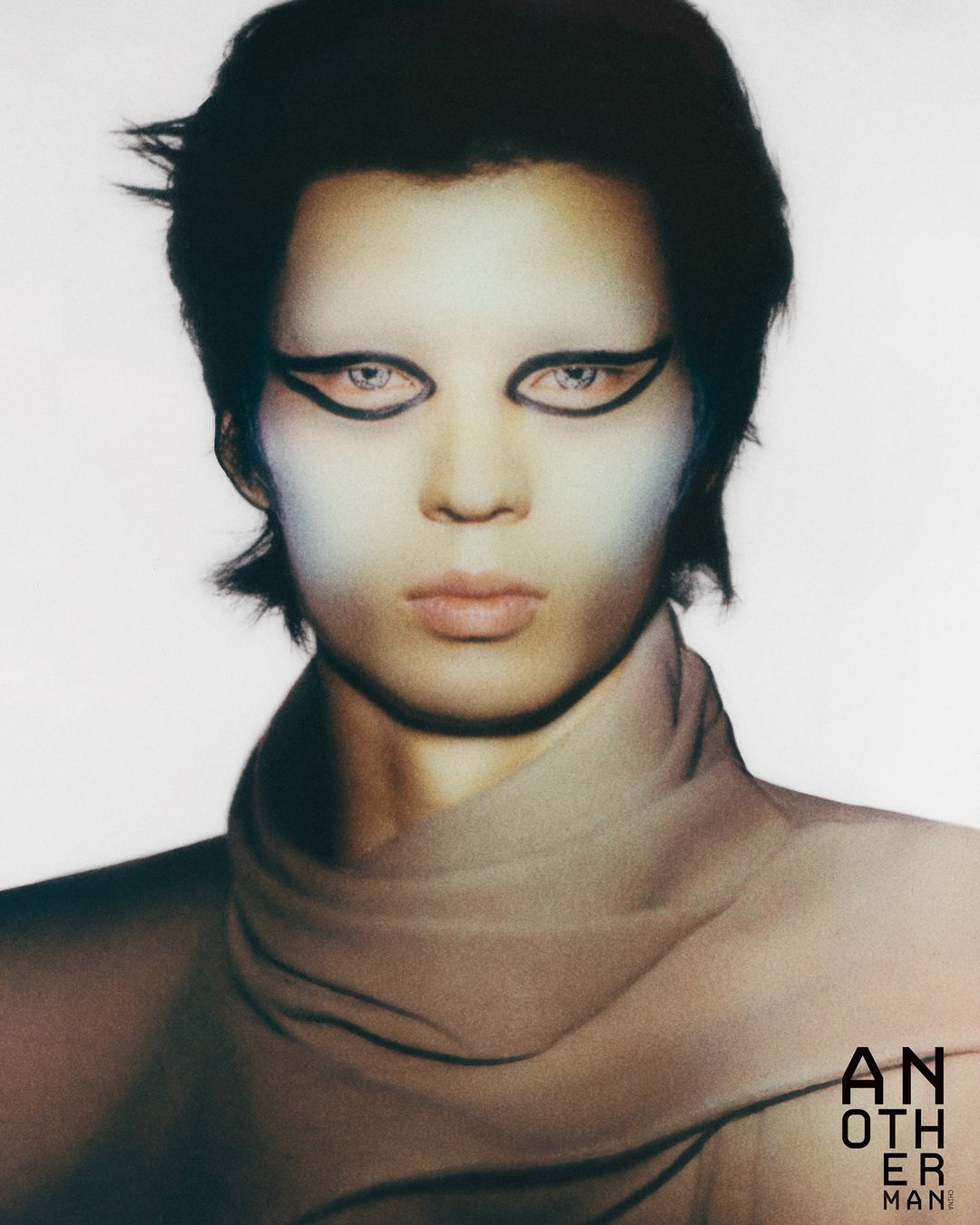 Close-up portrait of a person with dark hair and dramatic eye makeup, wearing a high-necked garment, against a light background.