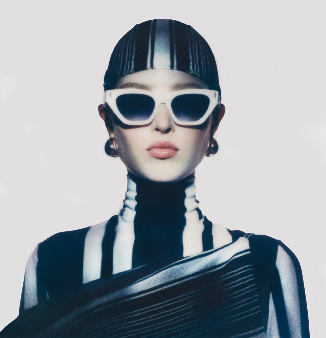 Model wearing white cat-eye sunglasses, a striped cap, and a matching turtleneck against a plain background