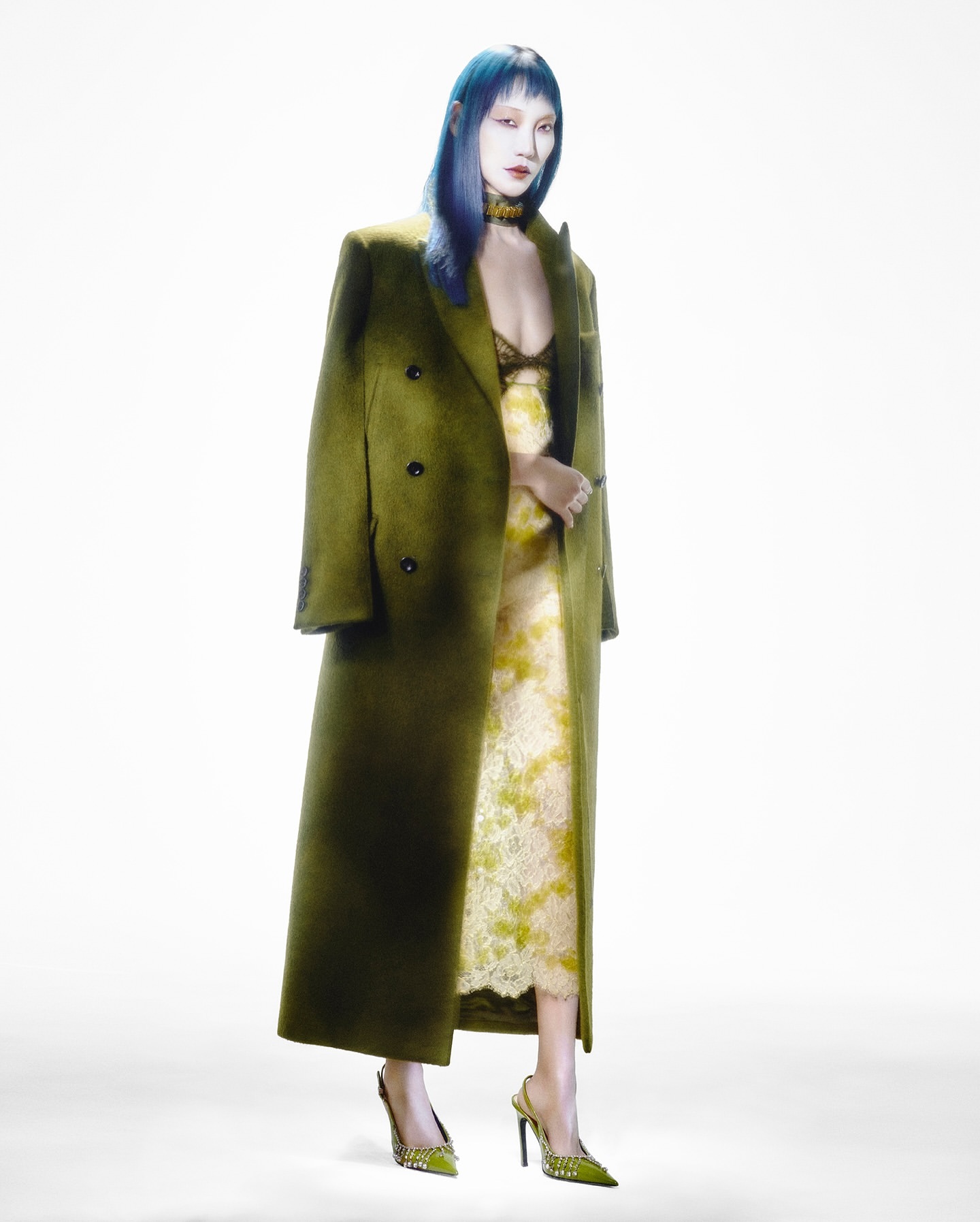 A woman with blue hair stands in a green coat and patterned dress in a fashion photography studio, posing against a clean white backdrop.