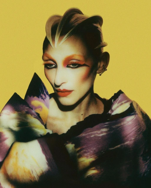 Portrait of a person with dramatic makeup and sculptural blonde hair, posed against a solid yellow backdrop, wearing colorful garments.