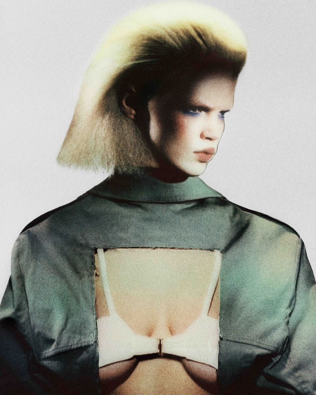 Fashion photo featuring a model with blonde hair and blue eye shadow wearing a collared shirt with a bra cut-out on a light background.