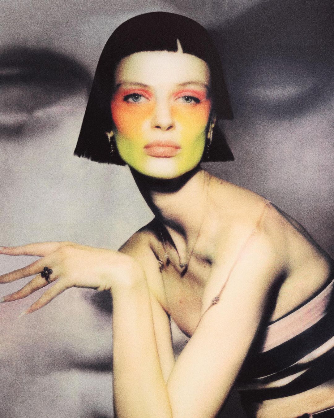 Close-up of a model with short black hair and orange, yellow, and green face paint, posing in front of a blurred gray background.