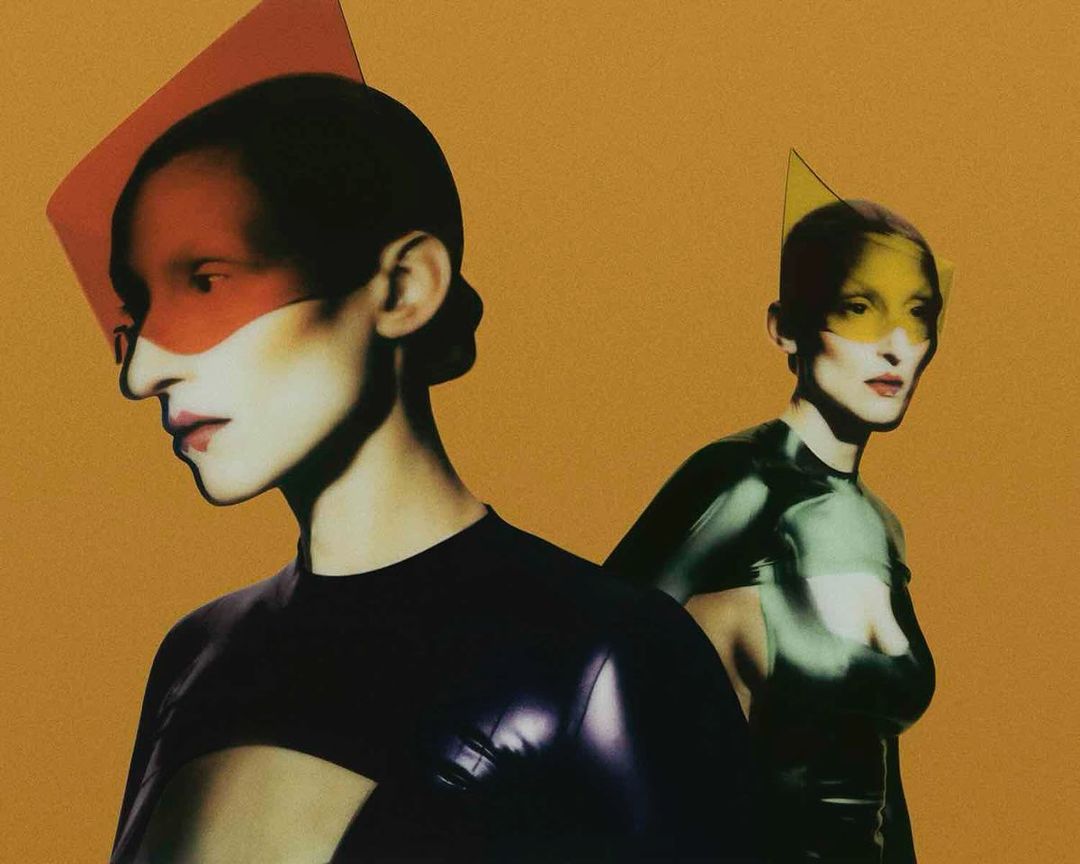 Two women in futuristic attire pose against an orange backdrop, faces partially obscured by geometric color filters, fashion-forward style.