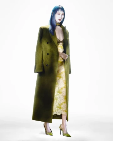 Model with vibrant blue hair wearing an olive green coat over a patterned dress against a white background.