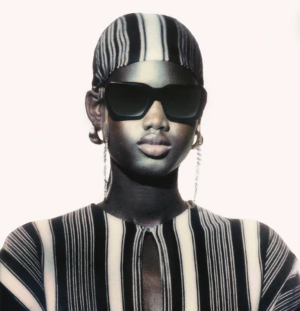 Close-up portrait of a Black woman wearing striped headwear, black sunglasses, and a matching striped top against a white backdrop.