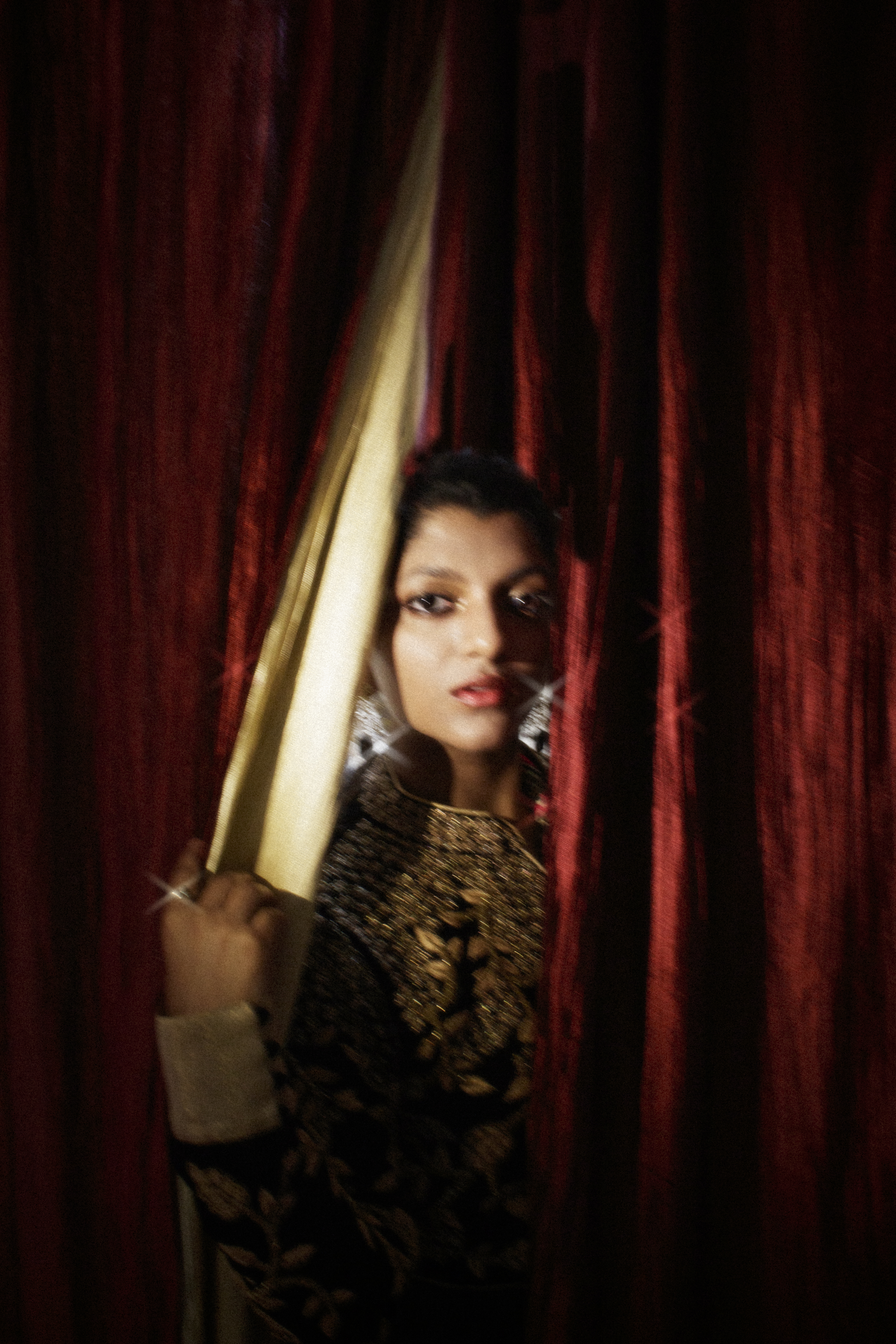 A woman peeking through a gap in heavy red curtains, wearing dark ornate embroidery and statement silver earrings.
