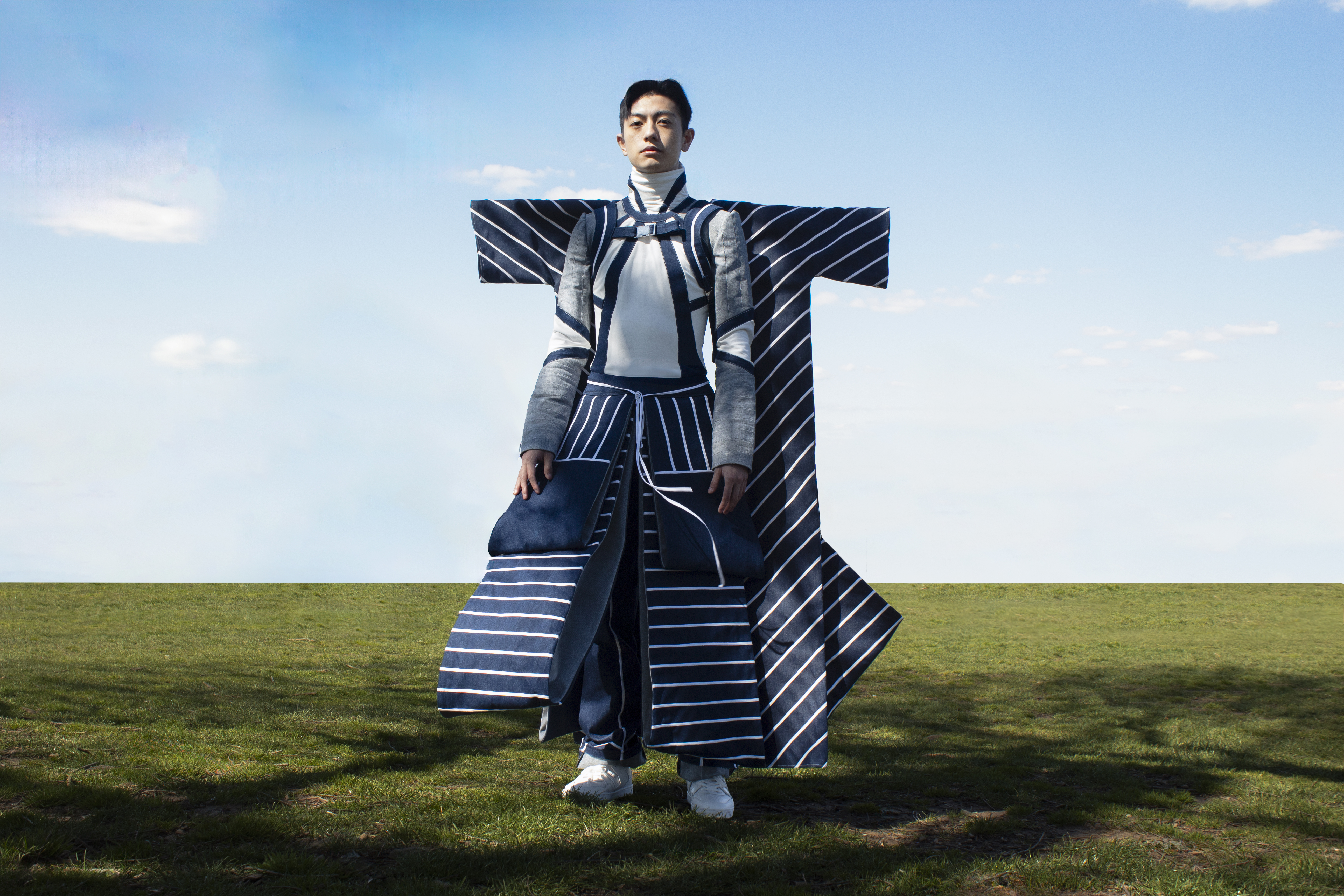 A man stands in a grassy field wearing a structured navy and white striped garment with exaggerated rectangular shoulder extensions.