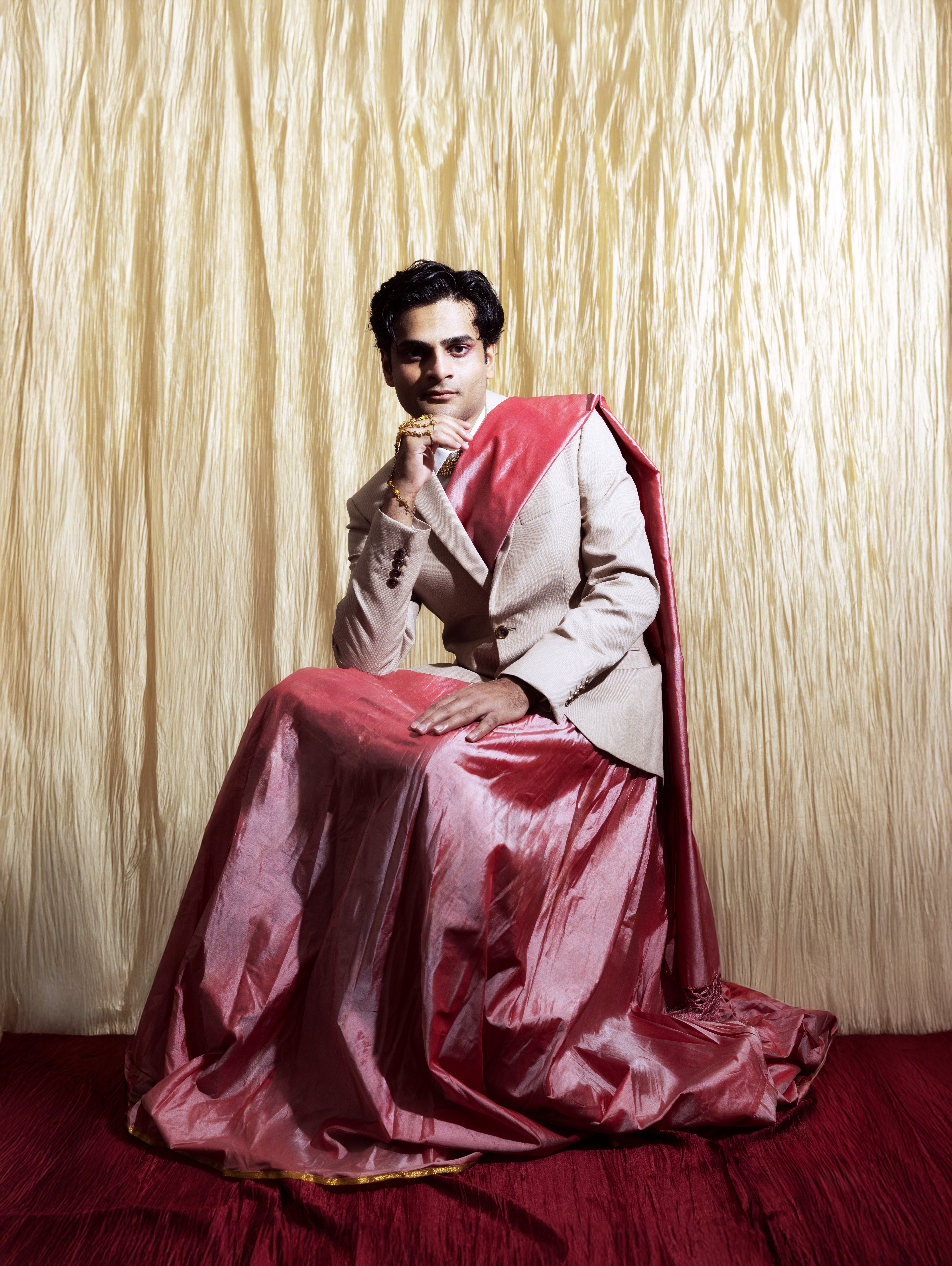 South Asian man wearing beige suit jacket and pink draped skirt seated against textured gold and red backdrop by Zoya RaZaiba