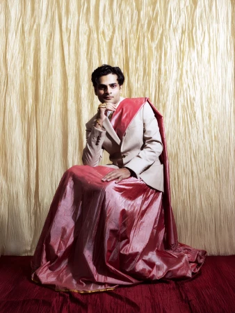 South Asian man wearing beige suit jacket and pink draped skirt seated against textured gold and red backdrop by Zoya RaZaiba