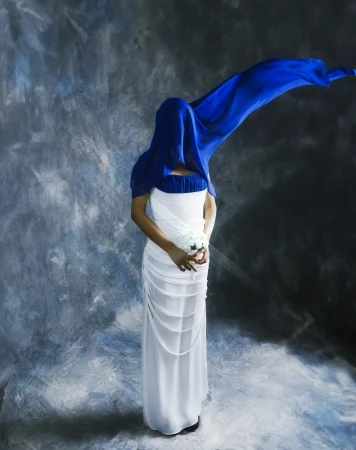 A person in a white wedding dress has their face obscured by a flowing royal blue veil against a textured grey studio backdrop.
