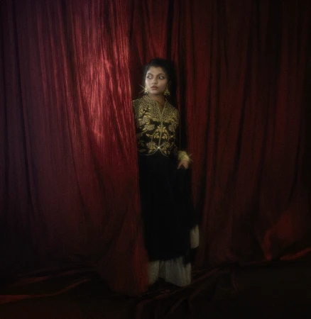 A woman wearing a dark black jacket with ornate gold floral embroidery stands partially hidden behind deep red velvet curtains.