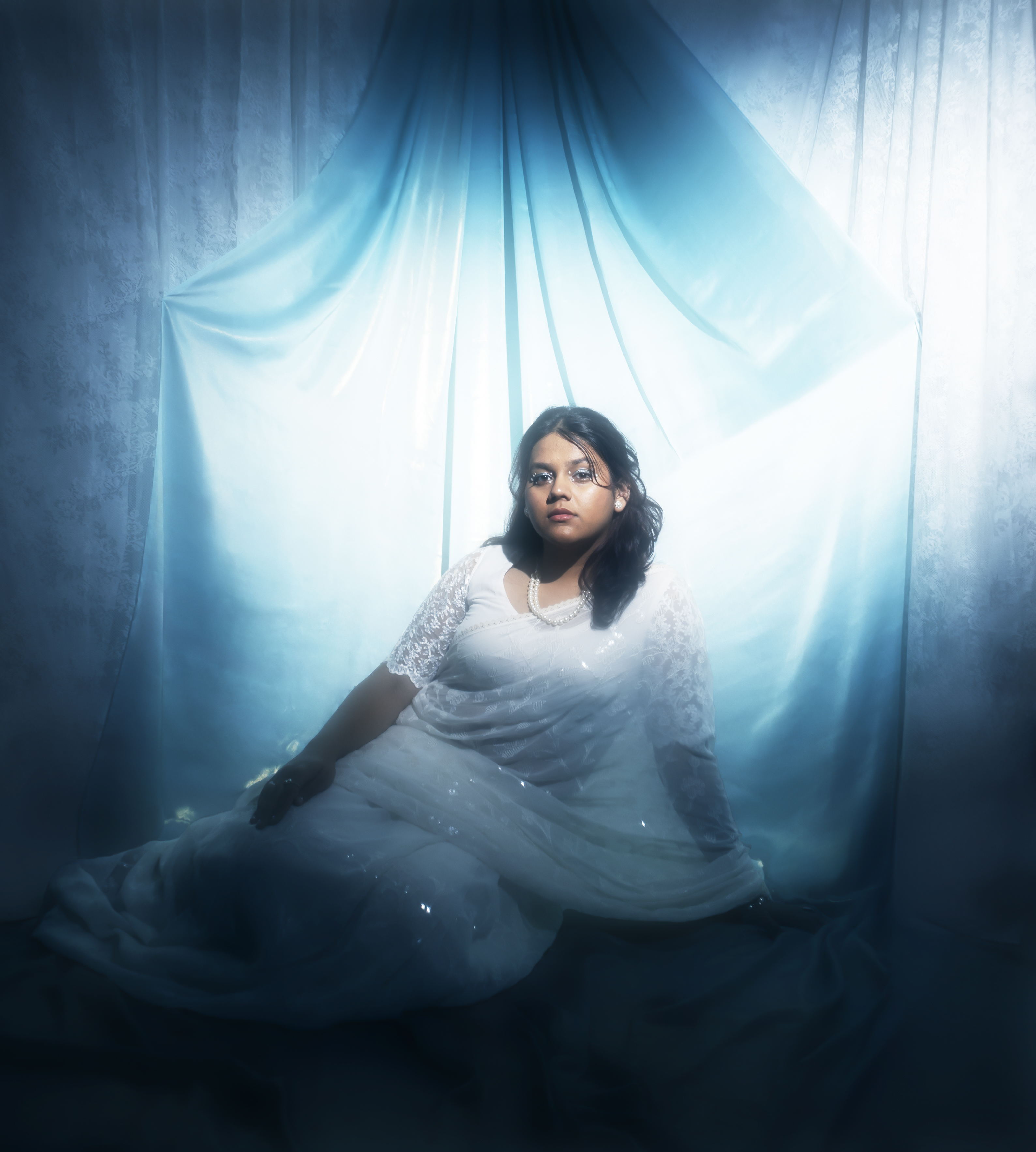 Woman in white lace dress sitting against a draped blue fabric backdrop with backlighting by Zoya RaZaiba