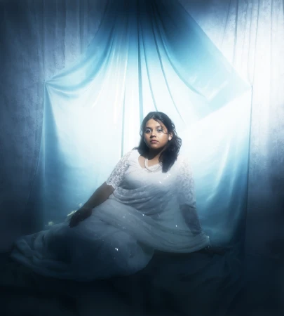 A woman in a white lace saree sits against a bright blue gradient fabric backdrop with soft cinematic lighting.