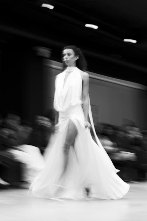 A model walks a runway in a flowing white halter dress with a high leg slit, captured with a motion-blurred effect in monochrome.