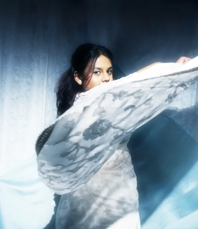 A young woman partially obscures her face with an intricate white embroidered lace fabric against a blue-toned textured backdrop.