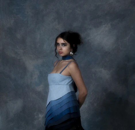 A young woman wearing a tiered light-to-dark blue ombre dress poses against a textured, moody blue-grey painted backdrop.