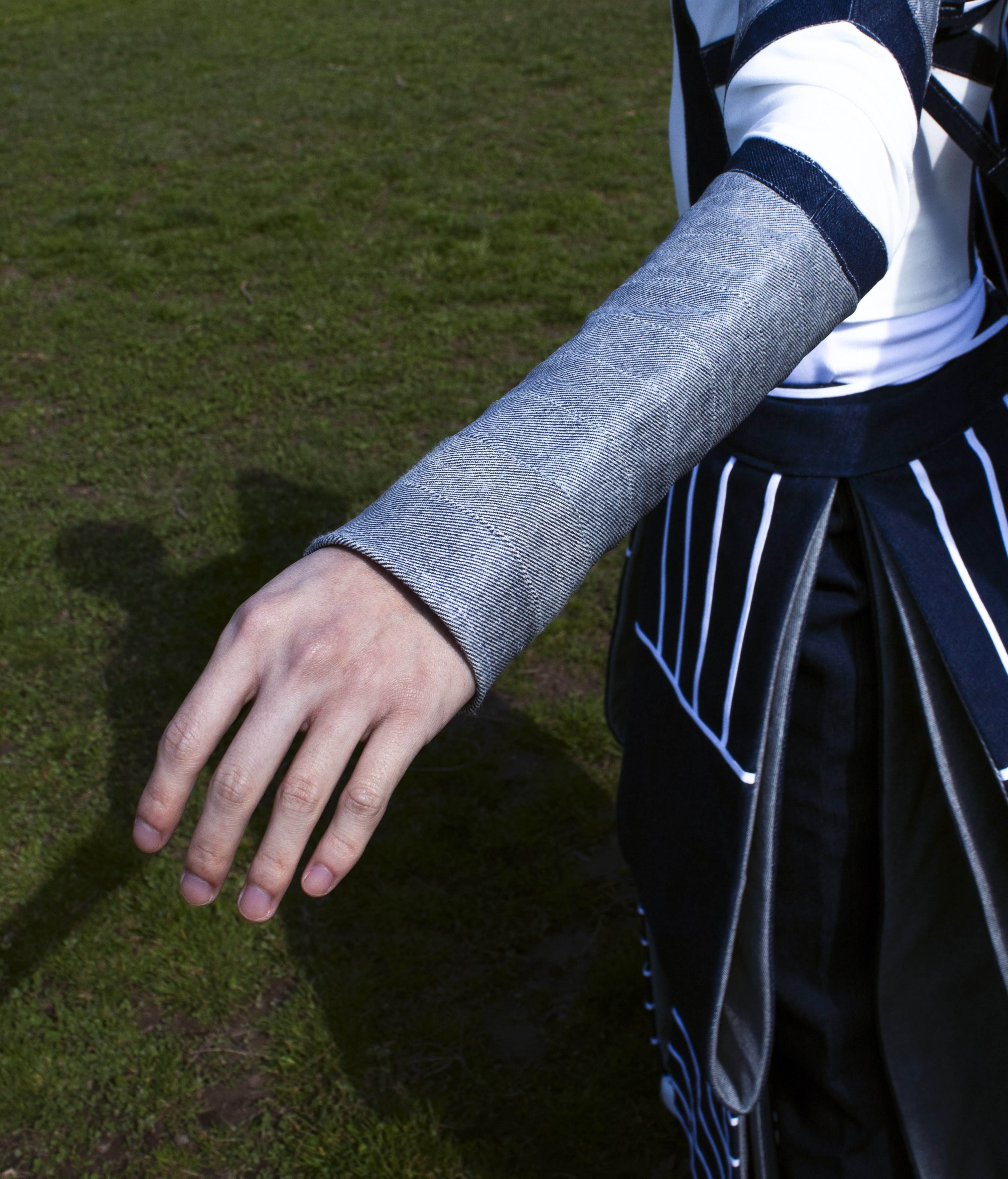 Close-up of a person's hand wearing a metallic silver fabric sleeve, paired with a black and white geometric skirt on a grassy field.