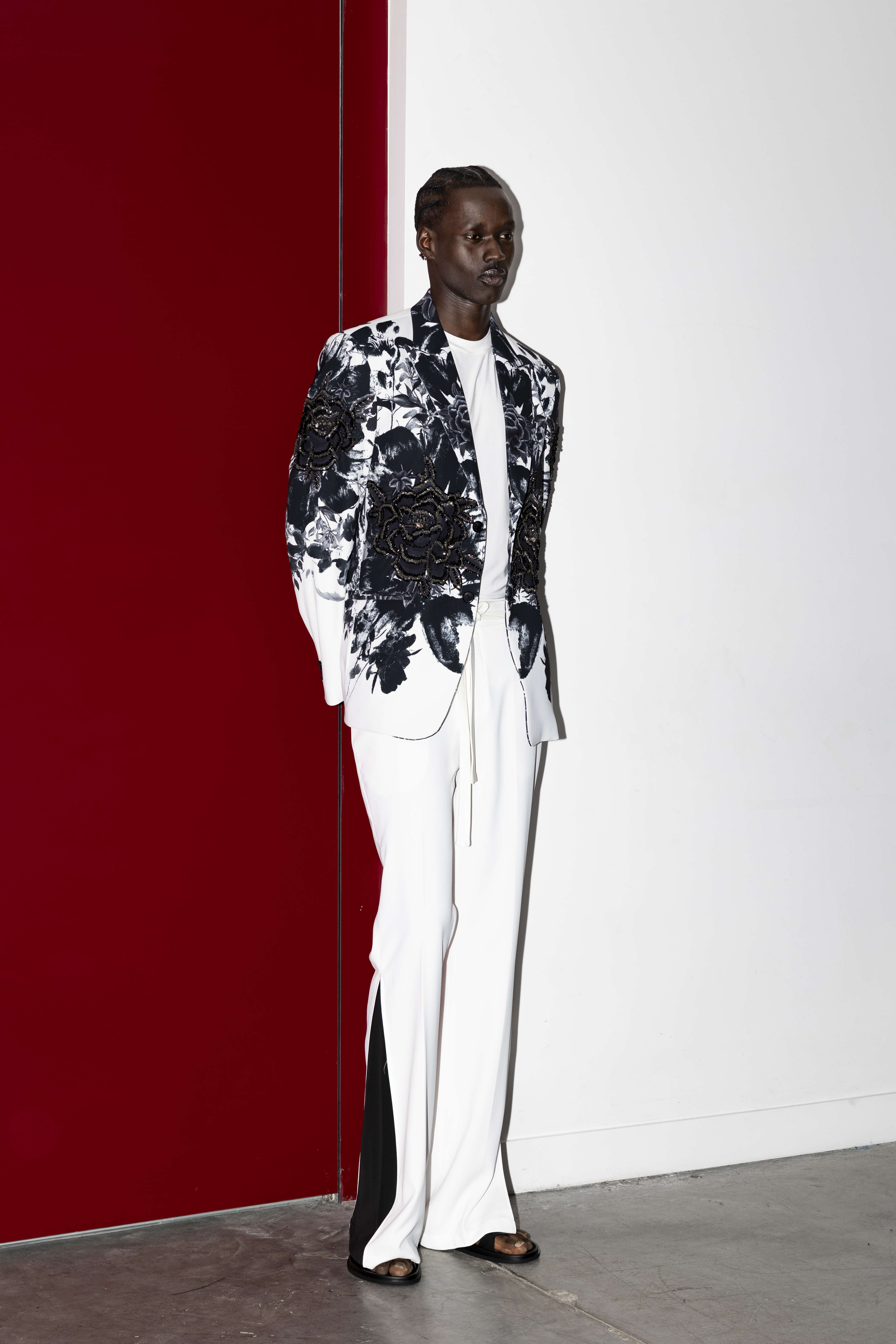 A man standing against a split red and white wall wearing a floral-print blazer and white trousers in a studio setting.