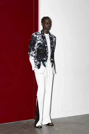 A man standing against a split red and white wall wearing a floral-print blazer and white trousers in a studio setting.