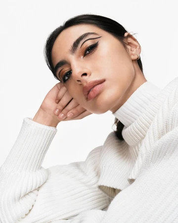 Portrait of a woman wearing a white ribbed turtleneck sweater with graphic eyeliner against a plain white background