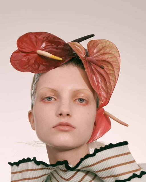 Close-up of woman with pale skin and anthurium flower headpiece, wearing a striped top against a pale neutral backdrop. Fashion editorial.