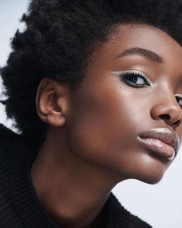 Close-up portrait of an African-American woman with natural hair, glossy lips, dramatic eyeliner and black top against a light background.