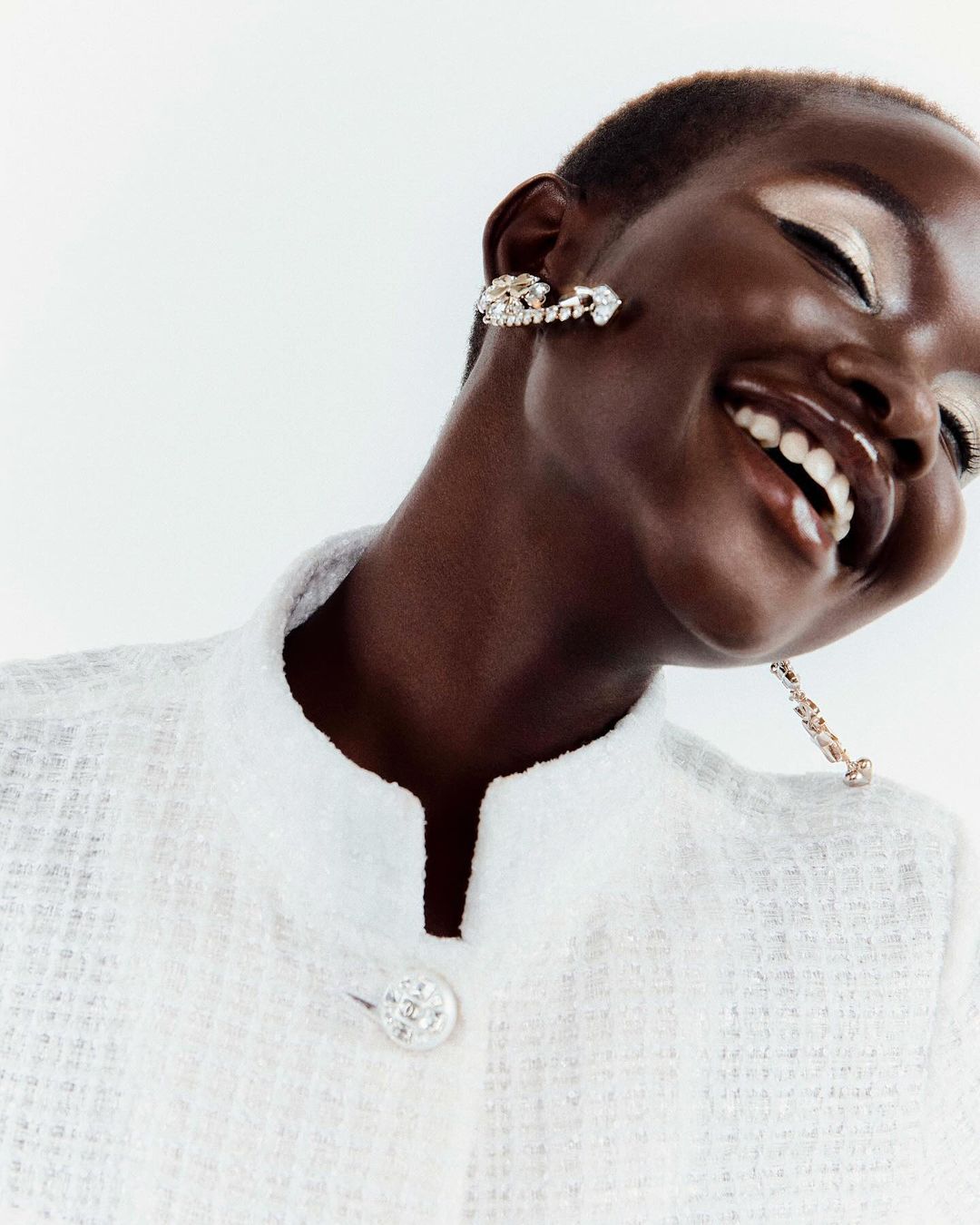 Close-up of a smiling Black woman with elegant earrings and shimmery eye makeup wearing a white tweed jacket against a white background.
