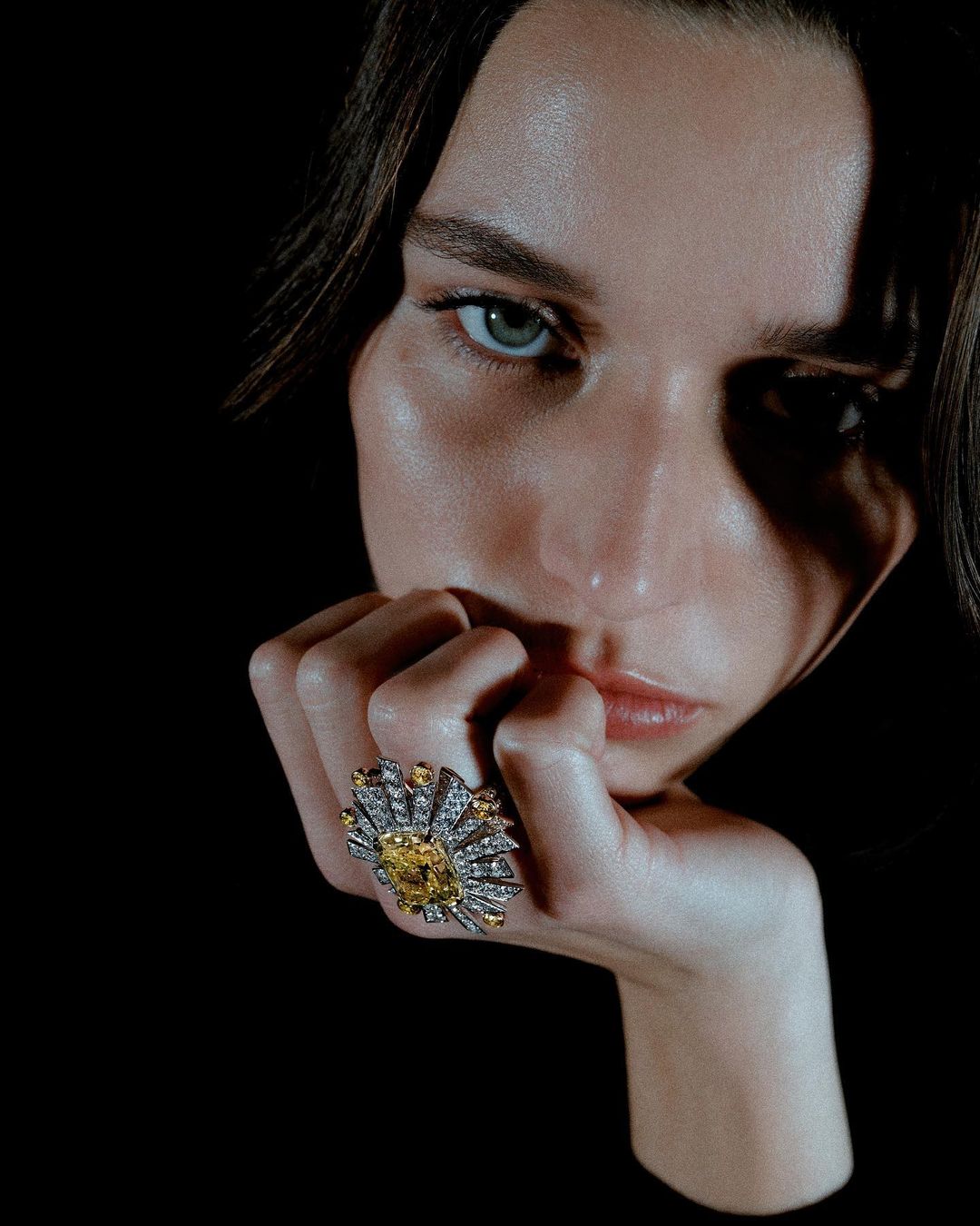 Close-up of a model with hand to face, showcasing a large, ornate yellow diamond ring against a stark black background.