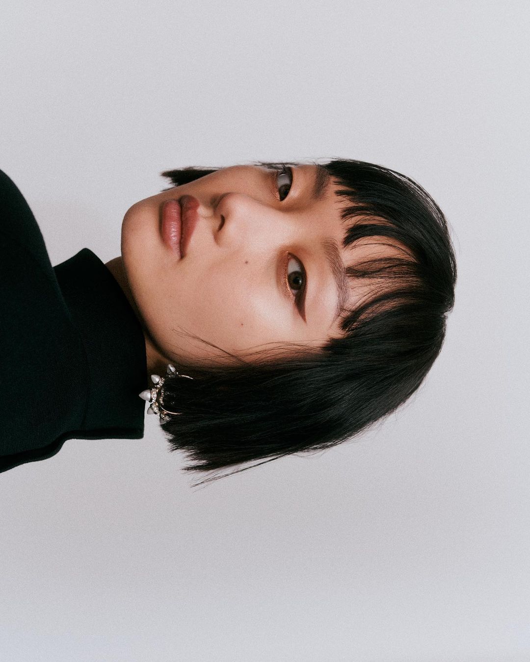 Portrait of an Asian woman with black short hair and blunt bangs wearing a black turtleneck and silver earring against a plain white background