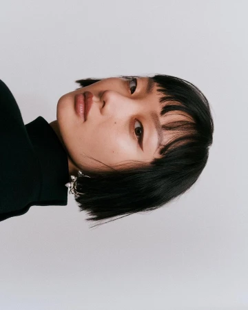Portrait of an Asian woman with black short hair and blunt bangs wearing a black turtleneck and silver earring against a plain white background