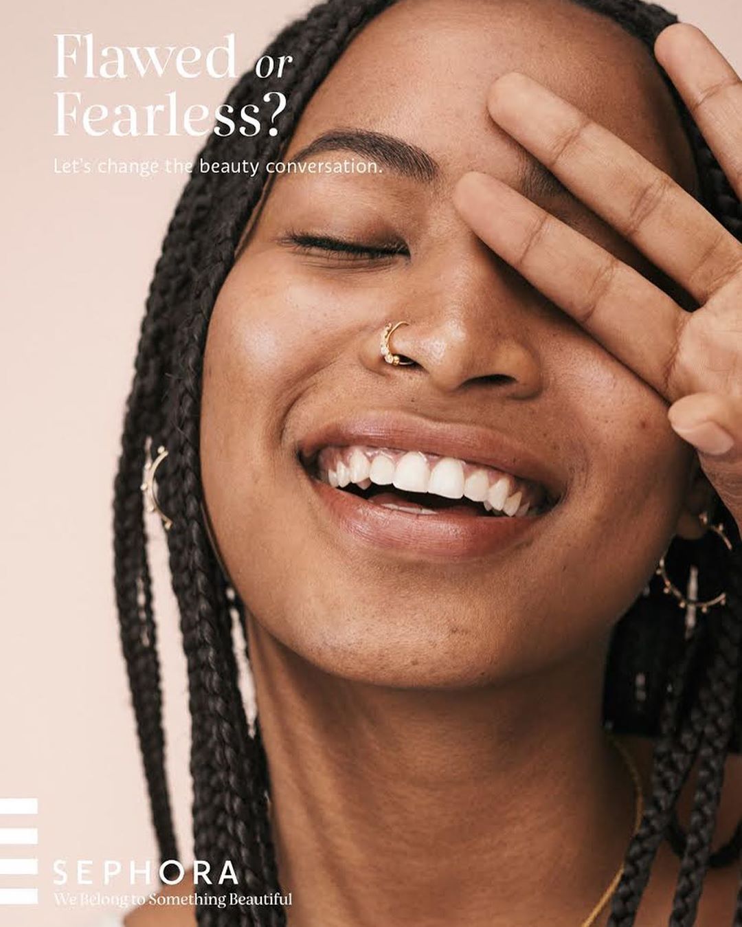Close-up of a smiling young Black woman with braided hair and a nose ring, covering one eye with her hand, in a beauty advertisement.
