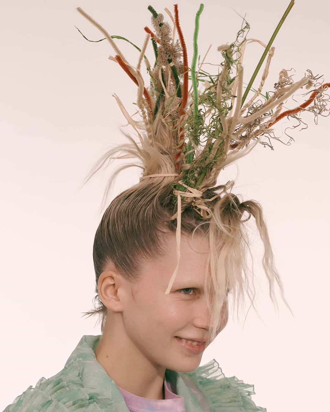 Close-up of smiling blonde woman with striking makeup and hair styled with decorative grass, posed in front of a bright background.