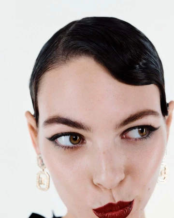 Close-up portrait of a woman with slicked-back dark hair, winged eyeliner, dark red lips, and gold geometric earrings against a white background.