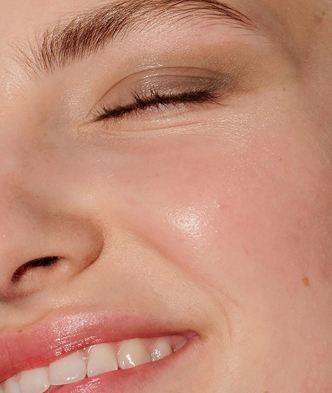 Close-up portrait of a smiling fair-skinned adult female with closed eyes, featuring clean makeup and glossy lips, in soft light.