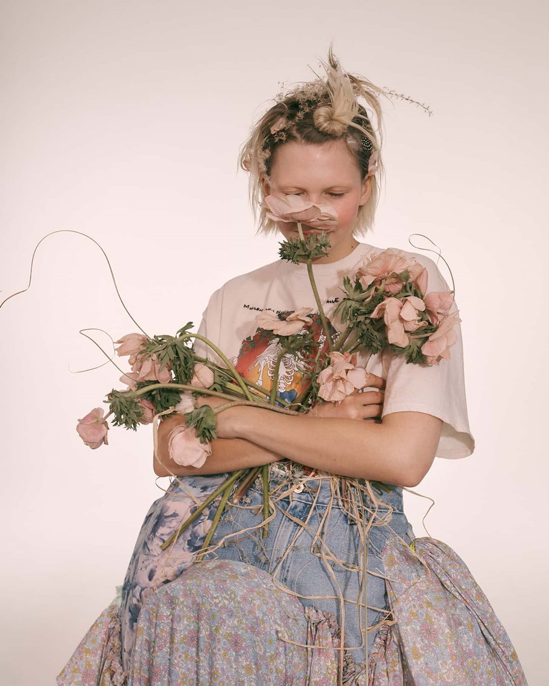 Young woman poses in a studio setting with a bouquet of pink flowers and a denim and floral dress. She's facing downwards in a portrait.