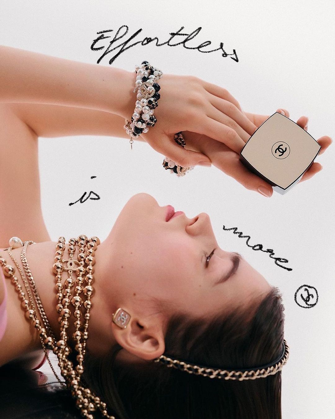 An adult female with light skin poses against a white background, accessorized with Chanel jewelry while holding a Chanel compact.