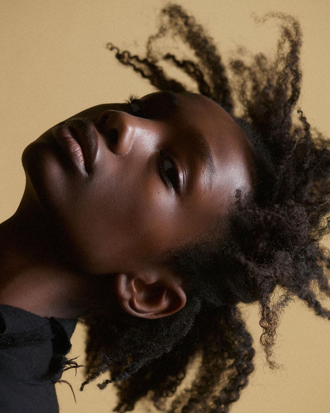 Close-up portrait of a Black woman with afro-textured hair against a light yellow background, showing her face and upper body.