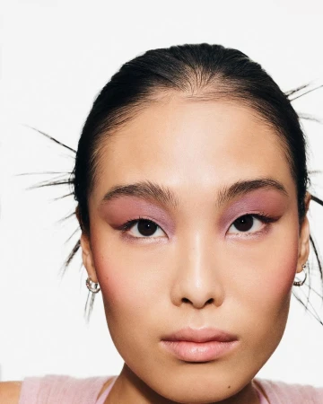 Close-up portrait of a model with pink eyeshadow and dark hair against a seamless white background.