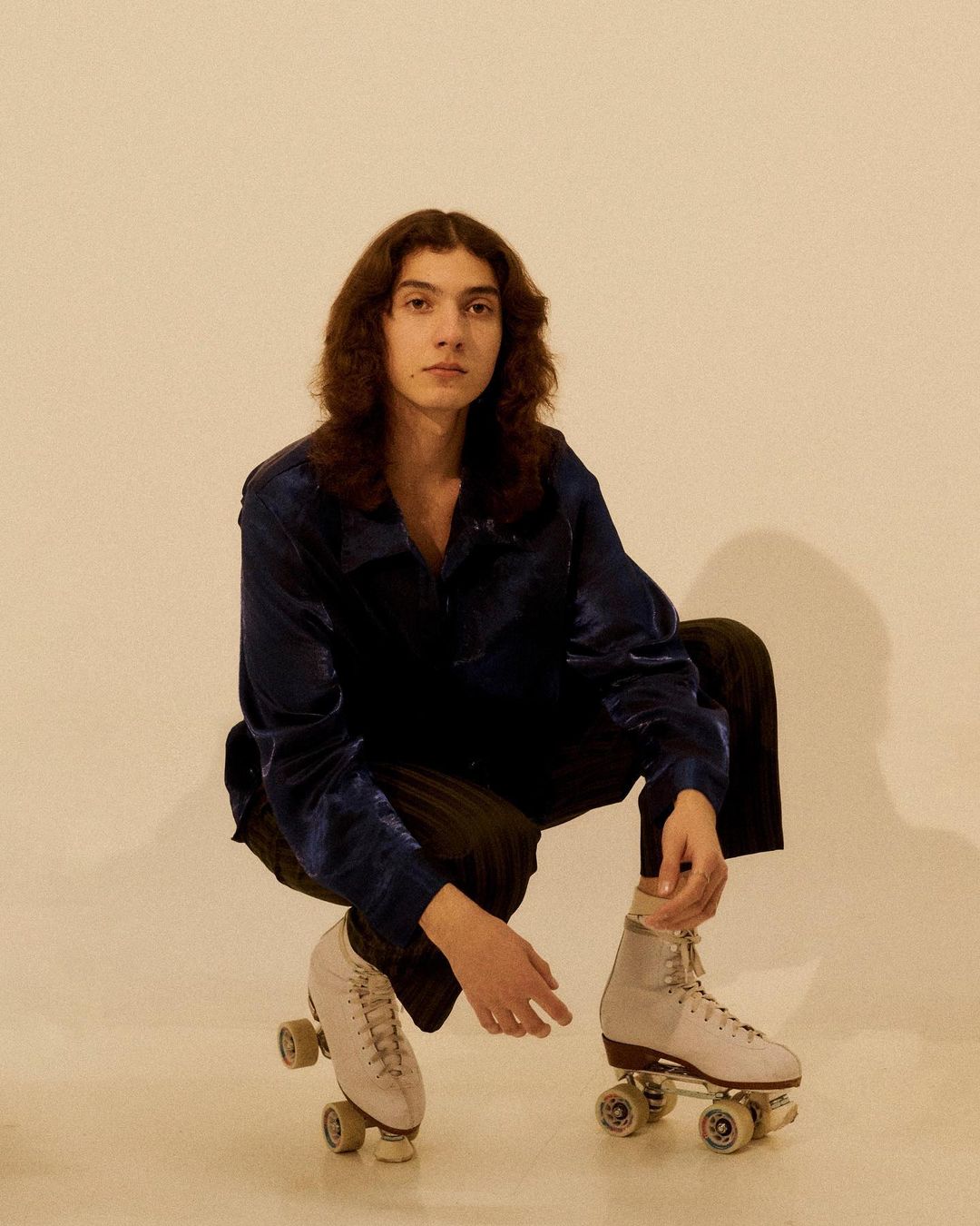 A young adult with long curly hair poses in a blue jacket and black pants, wearing roller skates in a studio shot on a cream background.