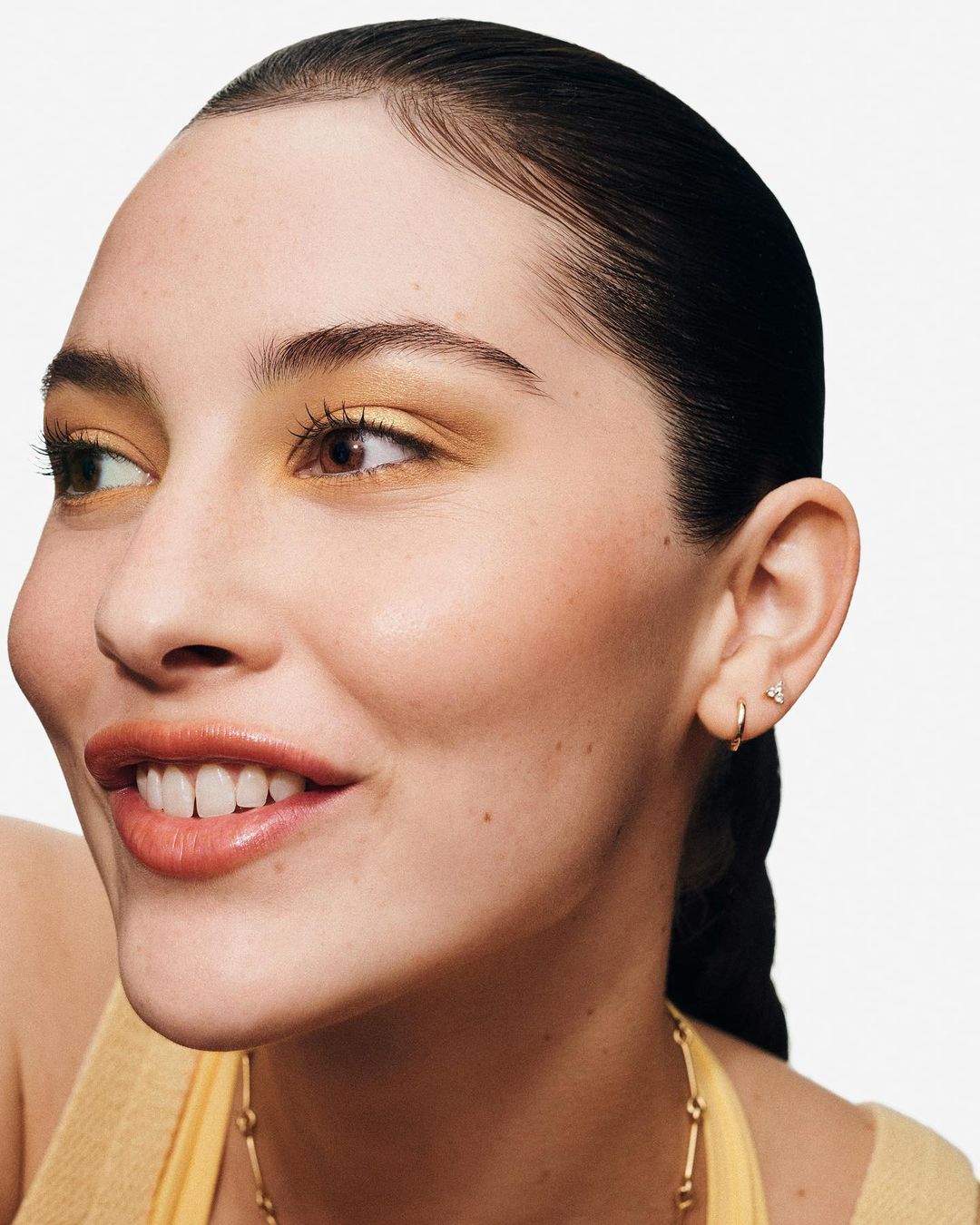 Close-up beauty shot of a smiling adult woman with yellow eyeshadow, gold necklace, and small gold earrings on a white background.
