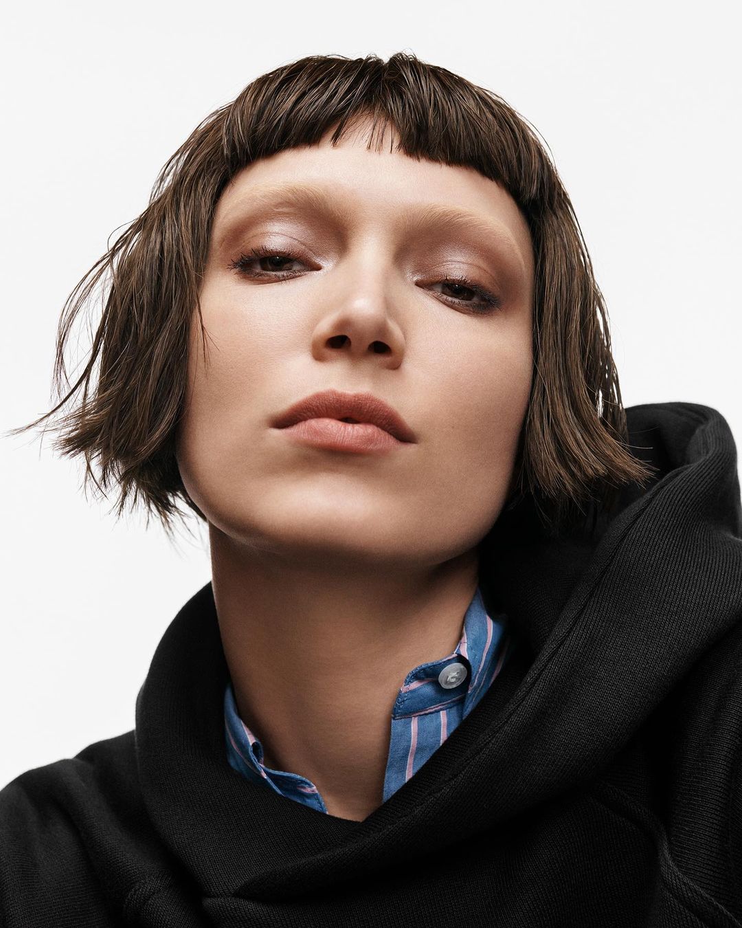 Close-up portrait of a fair-skinned adult woman with short brown hair and bangs, wearing a hooded jacket over a collared shirt in a studio.