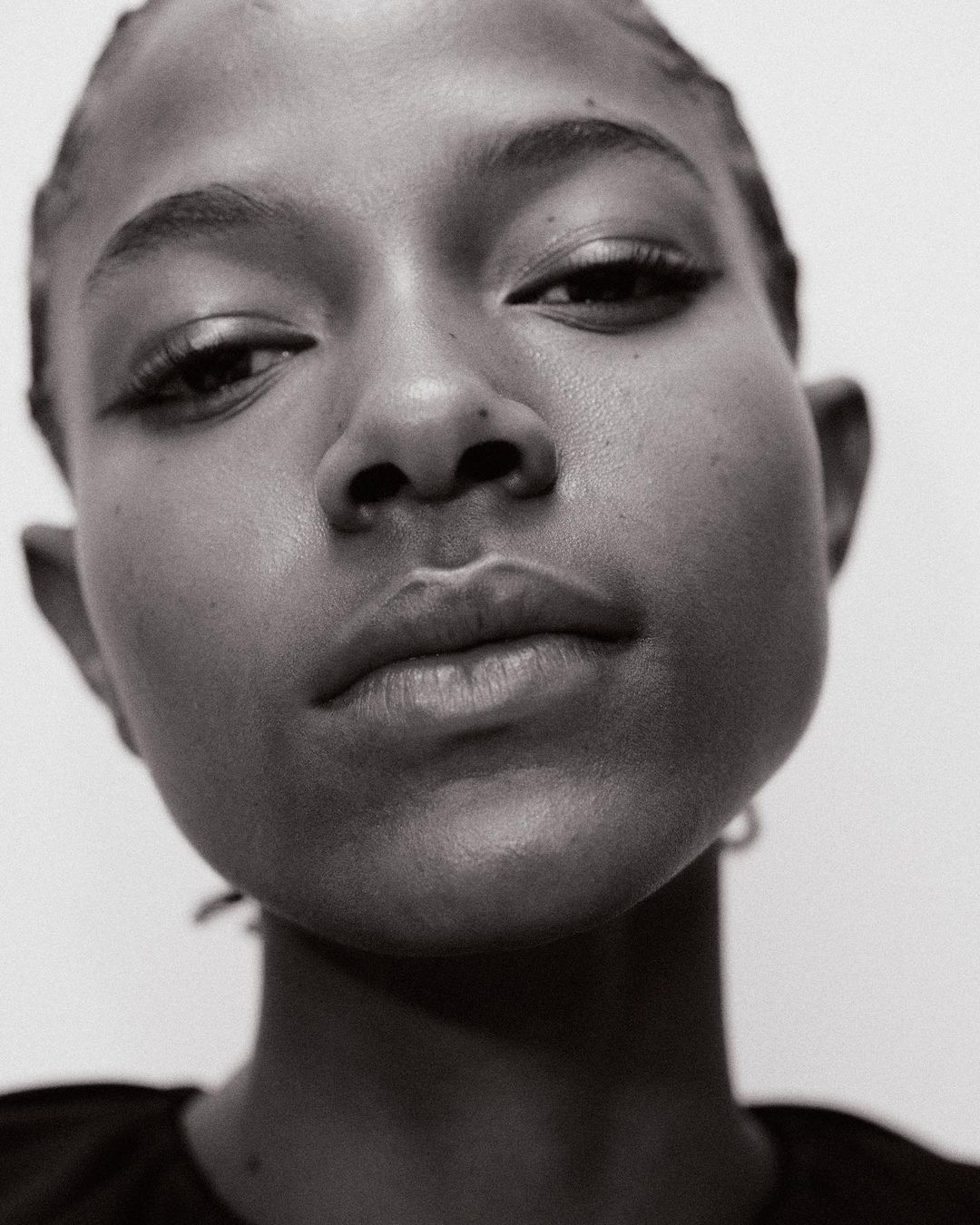 Close-up black and white portrait of a young Black woman with elegant eyeliner looking down against a stark white background.