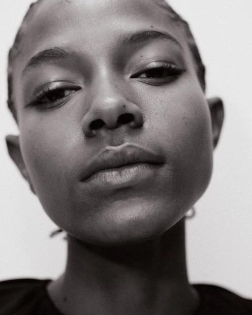 Close-up black and white portrait of a young Black woman with elegant eyeliner looking down against a stark white background.