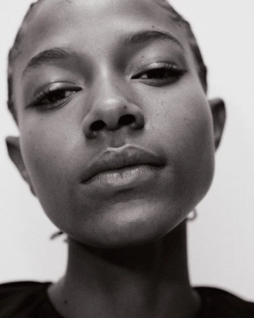 Close-up black and white portrait of a young Black woman with elegant eyeliner looking down against a stark white background.