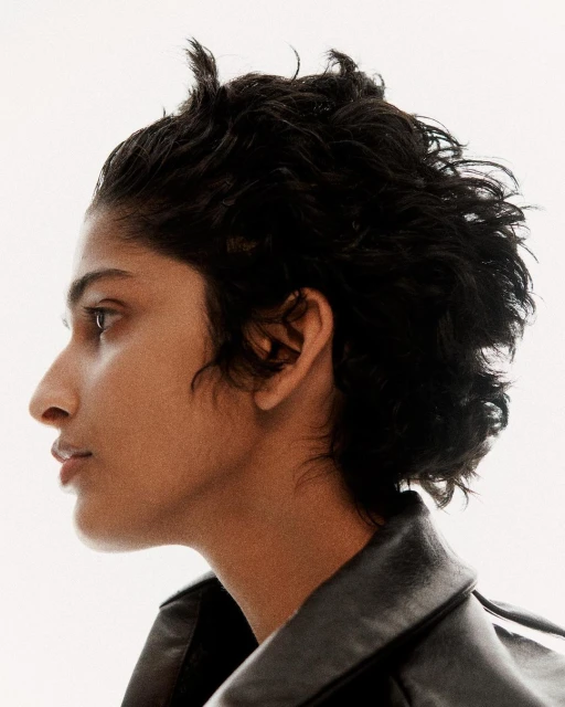 Profile portrait of a young adult woman with dark curly hair and a black leather jacket against a white background in a photo studio.