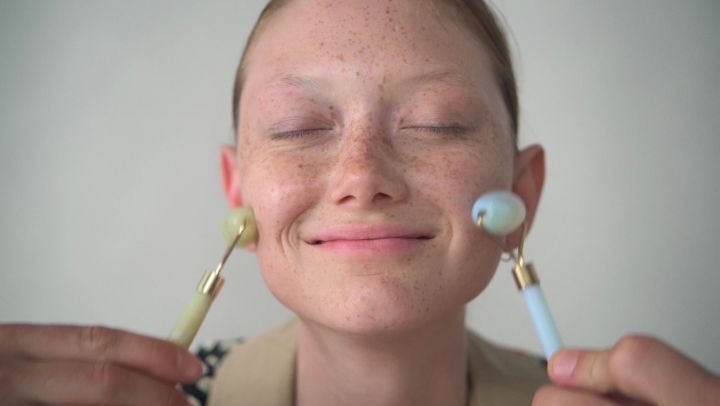 Close-up of a young woman with freckles using two facial massage rollers on her cheeks, indoors in front of a plain light background.